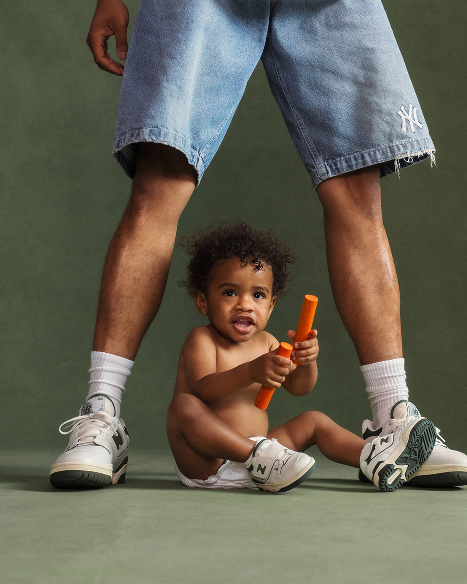 A photoshoot image of a man wearing shorts with a child between his legs